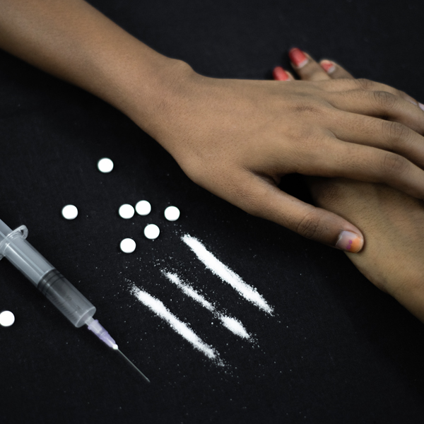 Hands holding each other next to drugs and a syringe on a black background, symbolizing support in the fight against addiction. It represents the process of Drug De-addiction, showing how unity helps overcome addiction.